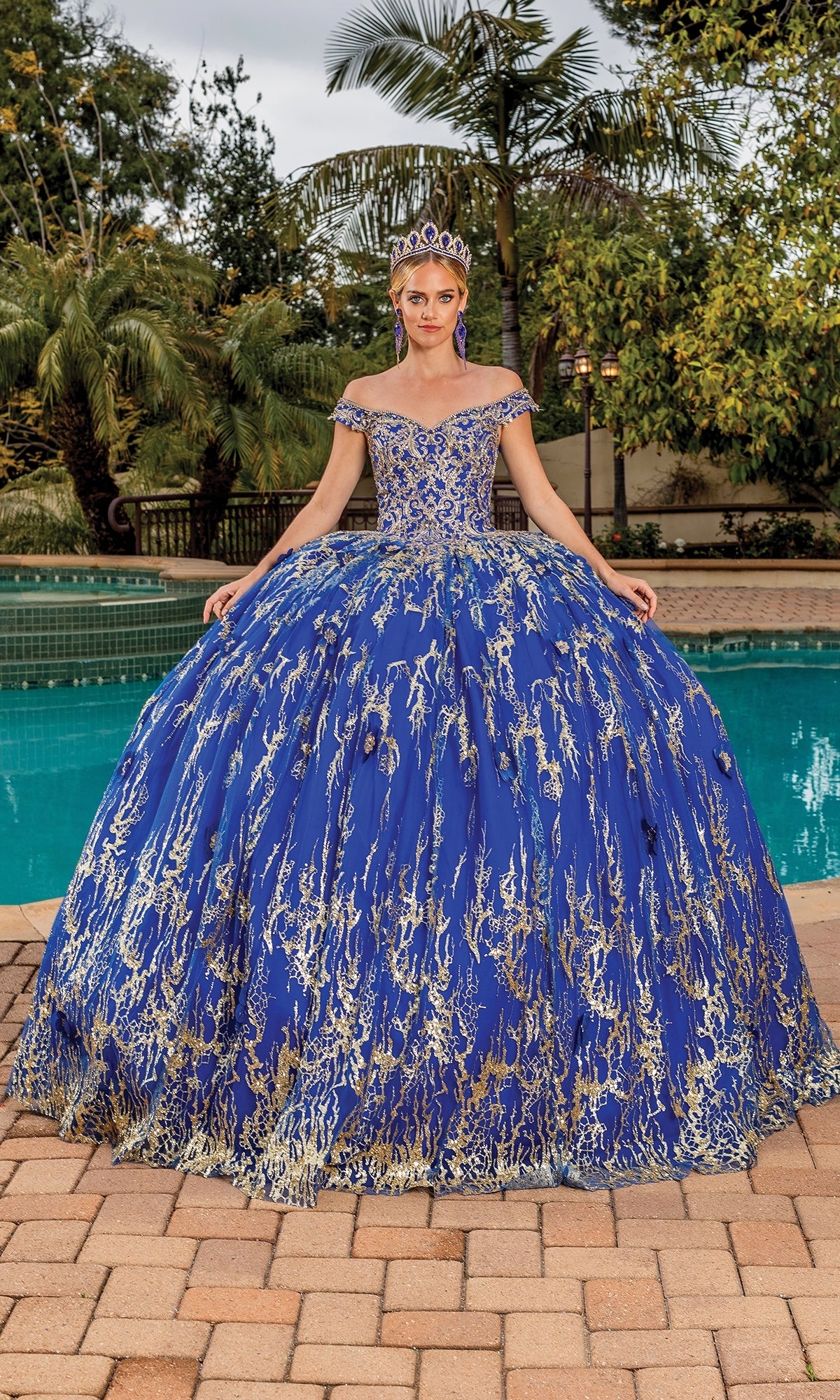 A woman in the Quinceanera Dress 1742 by Dancing Queen, featuring lace embroidery and off-the-shoulder straps, stands by a pool outdoors with greenery, wearing a matching tiara.